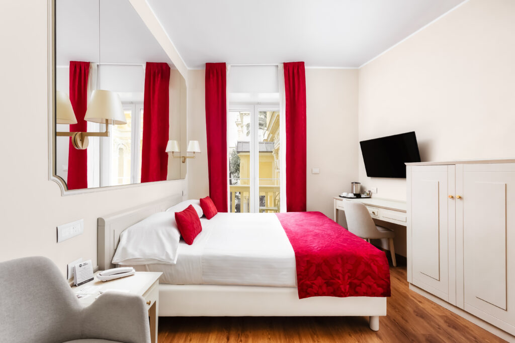 The image shows a double bedroom with French windows and a small balcony. Opposite is a desk and a wardrobe.