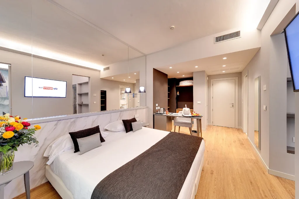 The image shows a suite with a window and a double bed. There is an open wardrobe and a desk.